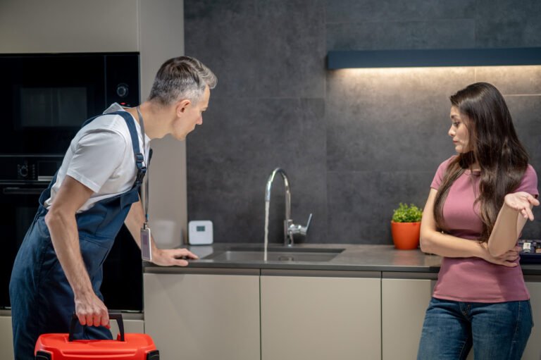 Breakdown, plumbing. Pretty dark haired middle aged woman in tshirt with question gesture and male plumber in overalls with special case looking at broken faucet in kitchen